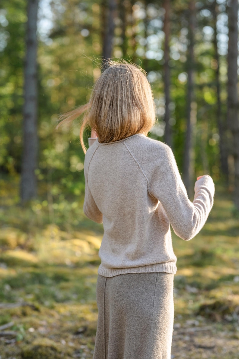 May include: A beige long-sleeved sweater with a round neckline and raglan sleeves, paired with a matching beige skirt. The sweater features a ribbed hem and cuffs, while the skirt has a visible vertical seam running down the center front.
