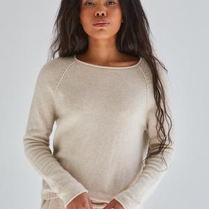 May include: A long-sleeved beige sweater featuring a round neckline with a subtle rolled edge, raglan sleeves, and a ribbed hem. The knit garment is crafted with a smooth texture and a consistent light tan color throughout the entire piece.