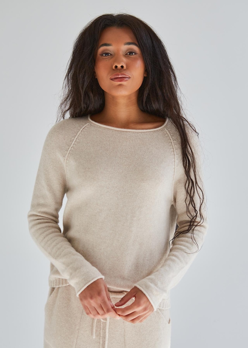 May include: A long-sleeved beige sweater featuring a round neckline with a subtle rolled edge, raglan sleeves, and a ribbed hem. The knit garment is crafted with a smooth texture and a consistent light tan color throughout the entire piece.