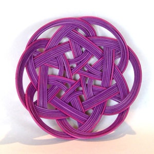 May include: A circular coaster with a complex, woven design. The coaster is predominantly purple with pink highlights, creating a striking contrast. The intricate pattern suggests a handcrafted item, suitable for home decor.