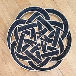 May include: A woven circular mat with a black and white design. The mat is made of thin strips of material that are woven together to create a complex pattern.