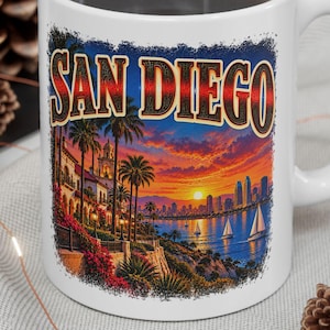 Könnte beinhalten: Ein weißer Keramikbecher mit einem farbenfrohen Grafikdruck der Skyline von San Diego bei Sonnenuntergang. Das Design zeigt Palmen, Segelboote auf dem Wasser und den Schriftzug SAN DIEGO in fetten, stilisierten Buchstaben über der Küstenstadt.