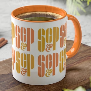 Puede incluir: Una taza de cerámica blanca con interior y asa naranja, llena de café. La taza presenta la palabra "SEP" en una fuente retro naranja y amarilla repetida. La taza está sobre una tabla de cortar de madera con palitos de canela y piñas.