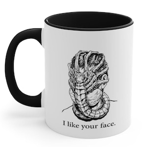May include: White ceramic mug with a black handle and interior. The mug features a black and white illustration of a facehugger from the Alien film franchise, with the text "I like your face."
