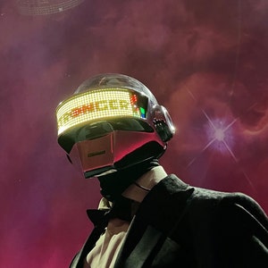 May include: A person wearing a silver and red helmet with the word "SINGER" displayed in green lights. The person is wearing a black suit and bow tie. The background is a dark purple and pink nebula.