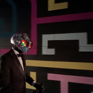 May include: A person wearing a silver helmet with a colorful visor and a black tuxedo with a bow tie. The background features a black wall with a colorful geometric design.