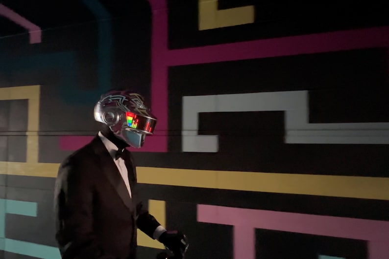 May include: A person wearing a silver helmet with a colorful visor and a black tuxedo with a bow tie. The background features a black wall with a colorful geometric design.