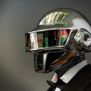 May include: A chrome-finished helmet with a visor displaying a colorful graphic equalizer. The helmet is part of a costume, worn with a black tuxedo and bow tie. The design is futuristic and reminiscent of electronic music artists.