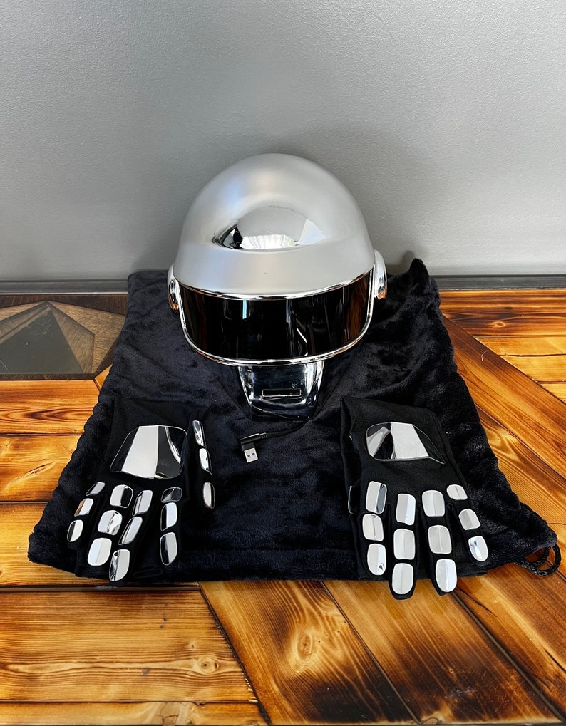 May include: A silver chrome helmet with a black visor, accompanied by a pair of black gloves with reflective silver accents. The items are displayed on a black velvet bag, suggesting a costume or performance theme.