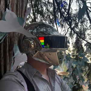 May include: A chrome-colored helmet with a rectangular visor displaying a colorful graphic. The helmet has a smooth, reflective surface and is worn by a person. The background includes trees and decorative paper cutouts.