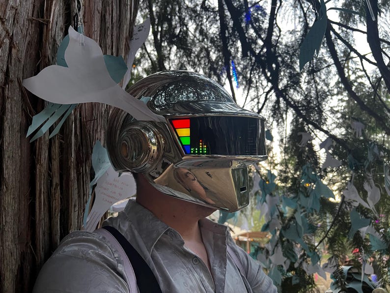 May include: A chrome-colored helmet with a rectangular visor displaying a colorful graphic. The helmet has a smooth, reflective surface and is worn by a person. The background includes trees and decorative paper cutouts.