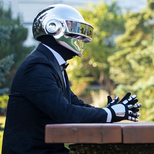 May include: A person wearing a chrome helmet with a digital display, black suit, bow tie, and gloves with chrome accents. The person is sitting at a brown table, with a green and yellow background.