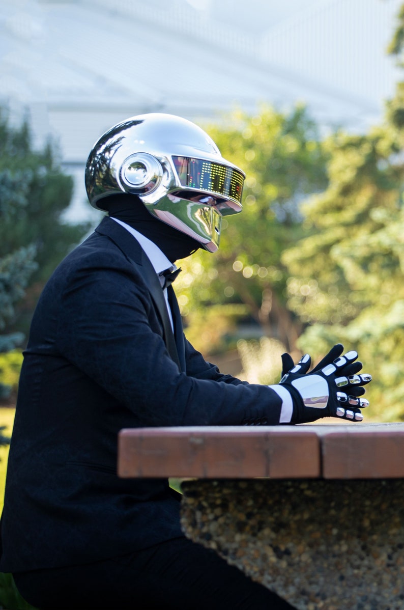 May include: A person wearing a chrome helmet with a digital display, black suit, bow tie, and gloves with chrome accents. The person is sitting at a brown table, with a green and yellow background.