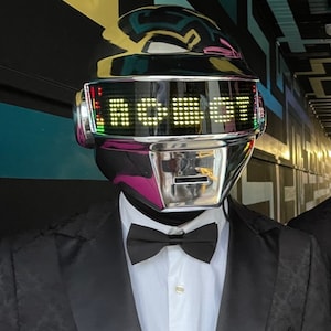 May include: A chrome robot helmet with the word "ROBOT" displayed in yellow lights. The helmet is worn over a white collared shirt, black bow tie, and a black tuxedo jacket.