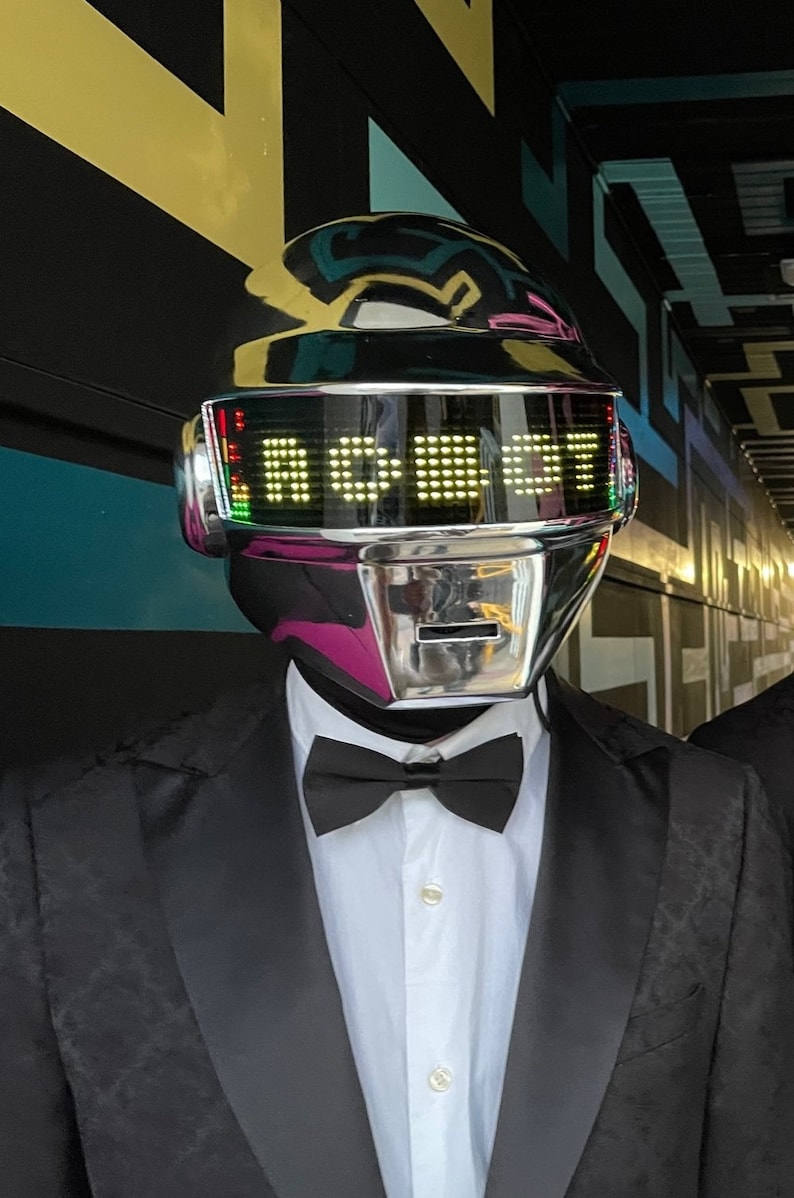 May include: A chrome robot helmet with the word "ROBOT" displayed in yellow lights. The helmet is worn over a white collared shirt, black bow tie, and a black tuxedo jacket.