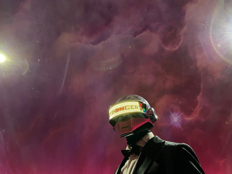 May include: A person wearing a silver and red helmet with the word "SINGER" displayed in green lights. The person is wearing a black suit and bow tie. The background is a dark purple and pink nebula.