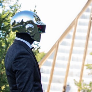 May include: A chrome-finished helmet with a rectangular display and a dark suit. The helmet reflects the surrounding environment. The suit includes a black bow tie and a white collared shirt. The background features greenery and a building.