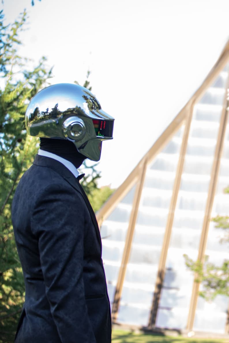 May include: A chrome-finished helmet with a rectangular display and a dark suit. The helmet reflects the surrounding environment. The suit includes a black bow tie and a white collared shirt. The background features greenery and a building.