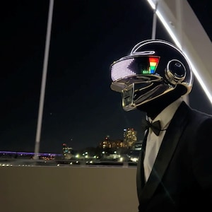 May include: A person wearing a black helmet with a reflective visor and a black suit with a bow tie. The helmet has a glowing LED display and a silver trim. The background shows a city at night.
