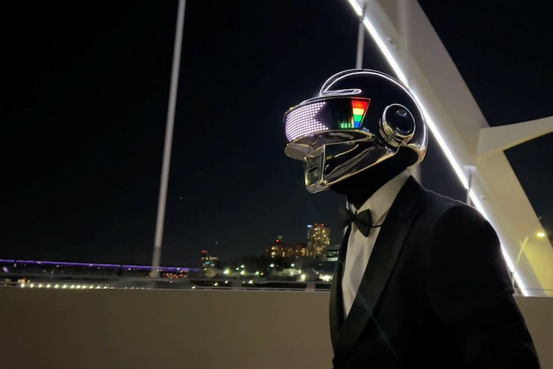 May include: A person wearing a black helmet with a reflective visor and a black suit with a bow tie. The helmet has a glowing LED display and a silver trim. The background shows a city at night.