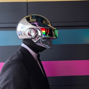 May include: A chrome-plated helmet with a rainbow display and a black neck covering. The helmet is worn with a black suit and bow tie. The background features black, gold, and pink stripes.