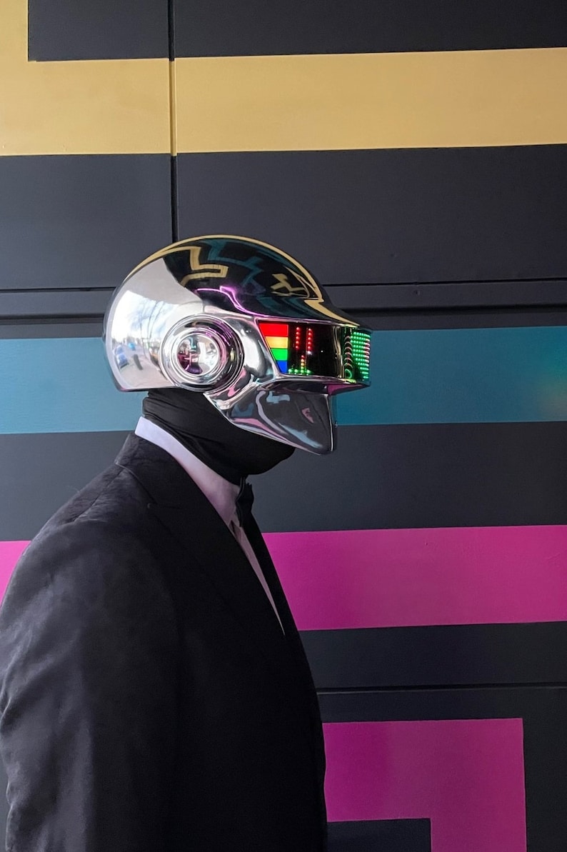 May include: A chrome-plated helmet with a rainbow display and a black neck covering. The helmet is worn with a black suit and bow tie. The background features black, gold, and pink stripes.