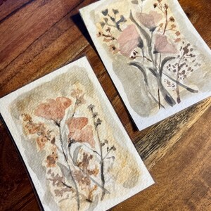May include: Two watercolor paintings of floral arrangements. The paintings feature soft pink and peach flowers with brown and green stems and leaves. The artwork is on rectangular paper with a beige background.