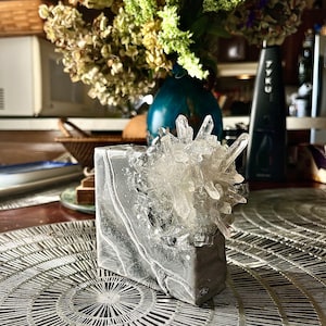 May include: A gray concrete block with a cluster of clear quartz crystals embedded in the top. The block is sitting on a gray circular placemat with a radial pattern.