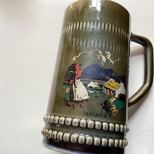 Puede incluir: Jarra de cerveza de cerámica verde oliva con asa, con una escena pintada de un paisaje irlandés con una figura con vestimenta tradicional, montañas y una cabaña. La palabra "Ireland" está impresa en la jarra.