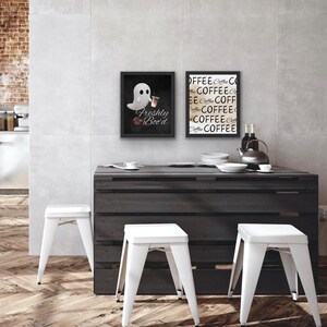 Puede incluir: Una escena de cocina con una mesa de madera oscura, taburetes blancos y arte enmarcado. Una impresión enmarcada dice "Freshly Boo'd" con un fantasma y café. La otra dice "Coffee" repetidamente. Una tetera y botellas están sobre la mesa.