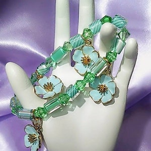 Może przedstawiać: Dwa zielone i niebieskie bransoletki z koralików z jasnoniebieskimi zawieszkami w kształcie kwiatów. Bransoletki znajdują się na białym stojaku w kształcie dłoni.