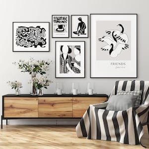 Könnte beinhalten: Eine Galeriewand mit gerahmten Schwarz-Weiß-Kunstdrucken. Die Drucke zeigen abstrakte Formen, Figuren und den Text "KAFFEEBREAK" und "FRIENDS forever". Ein Holzschrank, Blumen in Vasen und eine gestreifte Decke auf einem Stuhl vervollständigen die Szene.