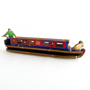 May include: A wooden model of a narrowboat with a red hull, blue trim, and a duck figurehead. The boat has several windows and a person standing on the deck.