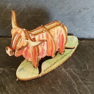 K&ouml;nnte beinhalten: Eine Holzfigur einer Highland Cow mit einem rot-orangen Streifenmuster. Die Figur steht auf einem Holzsockel mit dem aufgedruckten Text "www.etsy.com/shop/woodlandcraftsuk".