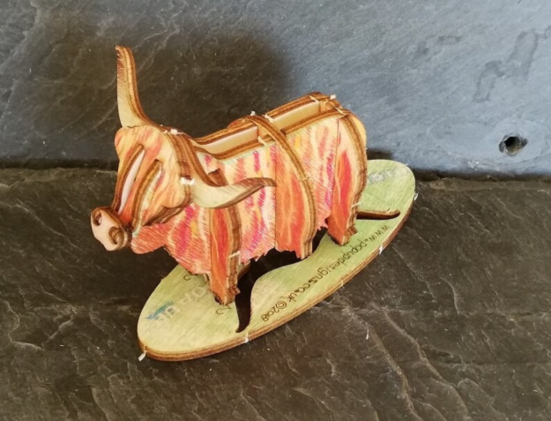 K&ouml;nnte beinhalten: Eine Holzfigur einer Highland Cow mit einem rot-orangen Streifenmuster. Die Figur steht auf einem Holzsockel mit dem aufgedruckten Text "www.etsy.com/shop/woodlandcraftsuk".