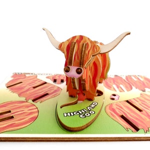 K&ouml;nnte beinhalten: Ein 3D-Puzzle aus Holz, das ein Highland Coo, eine schottische Rinderrasse, darstellt. Das Puzzle ist zusammengebaut und zeigt die Kuh auf einer gr&uuml;nen Weide stehend, mit den Worten "HIGHLAND COO" auf der Basis.