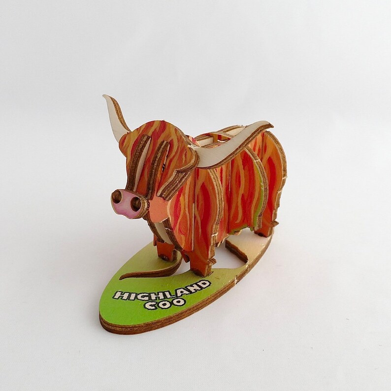 K&ouml;nnte beinhalten: Eine Holzfigur eines Highland Coo, einer schottischen Rinderrasse, mit einem rot-orangen gemusterten Fell. Die Figur steht auf einem gr&uuml;nen Sockel mit der Aufschrift "HIGHLAND COO".
