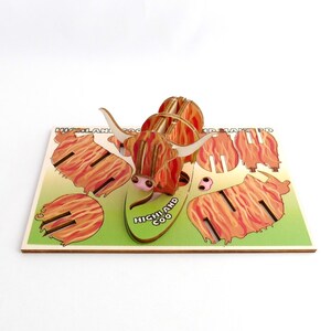 K&ouml;nnte beinhalten: Ein 3D-Puzzle aus Holz, das eine Highland-Kuh darstellt. Das Puzzle ist zusammengebaut und zeigt die Kuh auf einem gr&uuml;nen Hintergrund stehend, auf dem die Worte "HIGHLAND COO" aufgedruckt sind. Die Kuh ist braun und wei&szlig; mit einem rot-orangenen Muster.