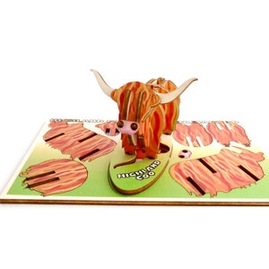 K&ouml;nnte beinhalten: Eine 3D-Pop-up-Karte mit einem Highland-Rind, einer schottischen Rinderrasse. Die Karte ist aus Holz und hat ein rot-orangenes Farbschema. Die Karte ist ge&ouml;ffnet und das Highland-Rind steht auf. Die Karte tr&auml;gt auch den Text "HIGHLAND COO".