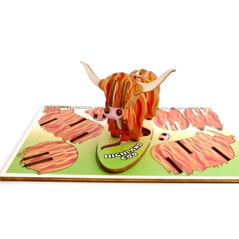 K&ouml;nnte beinhalten: Eine 3D-Pop-up-Karte mit einem Highland-Rind, einer schottischen Rinderrasse. Die Karte ist aus Holz und hat ein rot-orangenes Farbschema. Die Karte ist ge&ouml;ffnet und das Highland-Rind steht auf. Die Karte tr&auml;gt auch den Text "HIGHLAND COO".