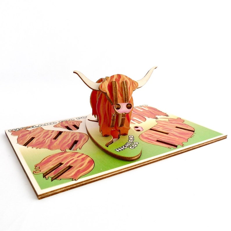 K&ouml;nnte beinhalten: Ein h&ouml;lzernes Highland Coo Spielzeug mit einem rot-braunen Streifenmuster. Das Spielzeug steht auf einem gr&uuml;n-braunen Sockel mit der Aufschrift "Highland Coo".