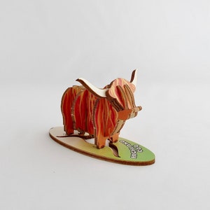 K&ouml;nnte beinhalten: Ein Holzmodell einer Highland-Kuh mit roten und orangen Streifen. Das Modell steht auf einem gr&uuml;nen Sockel mit der Aufschrift "Coo Highland".