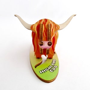 K&ouml;nnte beinhalten: Eine Holzfigur eines Highland Coo, einer schottischen Rinderrasse, mit einem rot-orangen Fellmuster. Die Figur steht auf einem gr&uuml;nen ovalen Sockel mit dem aufgedruckten Text "HIGHLAND COO".