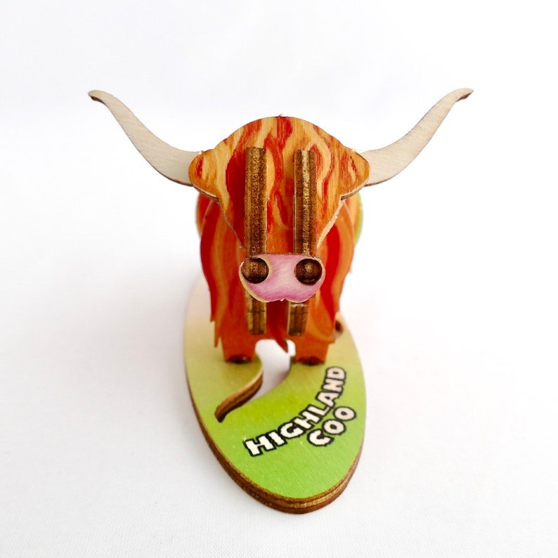K&ouml;nnte beinhalten: Eine Holzfigur eines Highland Coo, einer schottischen Rinderrasse, mit einem rot-orangen Fellmuster. Die Figur steht auf einem gr&uuml;nen ovalen Sockel mit dem aufgedruckten Text "HIGHLAND COO".