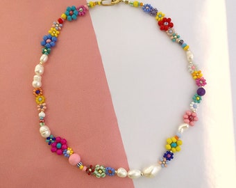 Può includere: Una collana di perline colorate con piccoli ciondoli a forma di fiore e accenti di perle. La collana è realizzata con una varietà di colori, tra cui rosa, blu, giallo, verde e rosso.