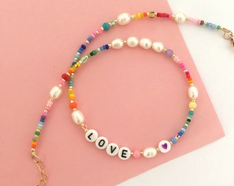 May include: A colorful beaded necklace with white pearls and the word "LOVE" spelled out in white beads. The necklace has a gold clasp.