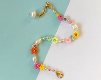 Può includere: Un braccialetto delicato con una catena e una chiusura, decorato con perline a forma di fiore colorate e perle bianche. Il braccialetto è un mix vibrante di rosa, arancio, giallo, blu e verde.