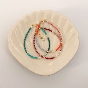 Könnte beinhalten: Vier farbige Perlenarmbänder mit goldenen Verschlüssen und Perlenakzenten. Die Armbänder sind in einem Kreis auf einer weißen Muschelform angeordnet.