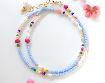 Può includere: Una delicata collana a due strati con perline di colore azzurro, bianco, rosa, giallo, verde e blu. La collana ha una chiusura dorata ed è impreziosita da piccole perle.