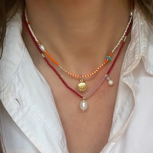 May include: A multi-strand necklace with beaded chains in red, orange, and pink. The necklace features a gold seashell pendant and several pearls. The chains are made of small beads, creating a delicate and layered look.